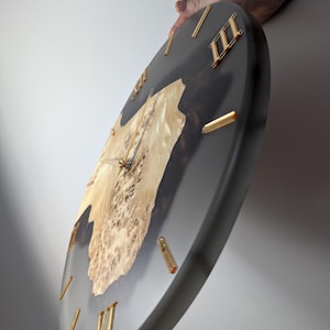May include: A round wall clock with a dark gray face and gold-colored Roman numerals. The clock features a natural wood burl center, adding a unique, organic element to the design. The hands are gold-colored.