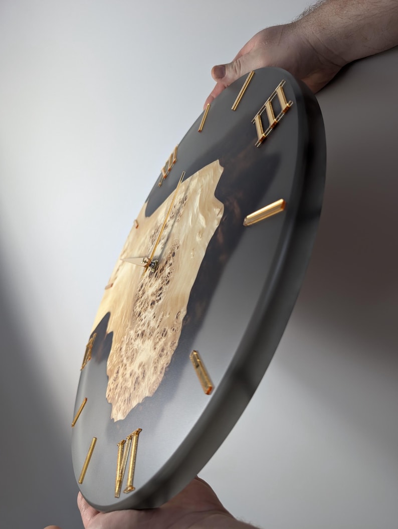May include: A round wall clock with a dark gray face and gold-colored Roman numerals. The clock features a natural wood burl center, adding a unique, organic element to the design. The hands are gold-colored.