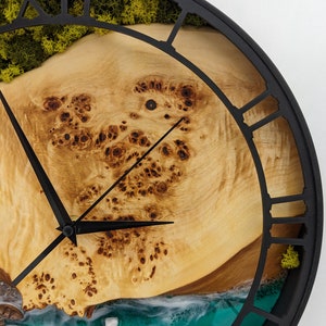 May include: A round wall clock with a black metal frame and Roman numerals. The clock face features a light brown wood grain design, with a turquoise and white ocean scene at the bottom. Green moss is visible at the top.