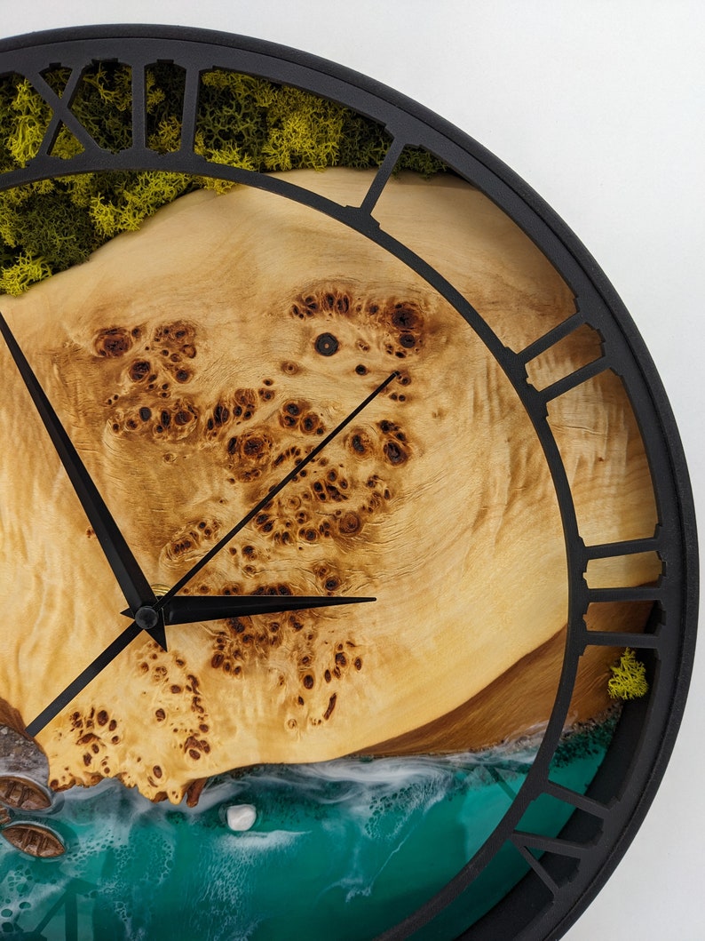 May include: A round wall clock with a black metal frame and Roman numerals. The clock face features a light brown wood grain design, with a turquoise and white ocean scene at the bottom. Green moss is visible at the top.