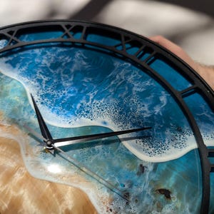 Pu&ograve; includere: Un orologio da parete rotondo con una cornice in metallo nero e numeri romani. Il quadrante presenta una scena oceanica dettagliata con acqua blu, schiuma bianca e una spiaggia sabbiosa. Le lancette dell'orologio sono nere.