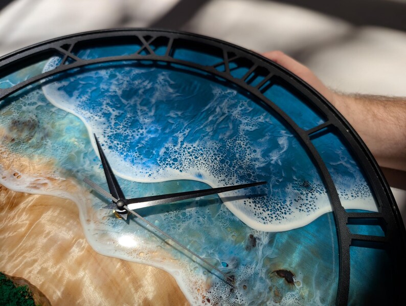 Pu&ograve; includere: Un orologio da parete rotondo con una cornice in metallo nero e numeri romani. Il quadrante presenta una scena oceanica dettagliata con acqua blu, schiuma bianca e una spiaggia sabbiosa. Le lancette dell'orologio sono nere.