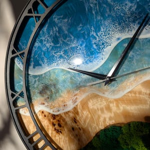 Pu&ograve; includere: Un orologio da parete rotondo con una cornice in metallo nero. Il quadrante dell'orologio presenta una scena di spiaggia con oceano blu, onde bianche e una riva sabbiosa. Le lancette dell'orologio sono nere.
