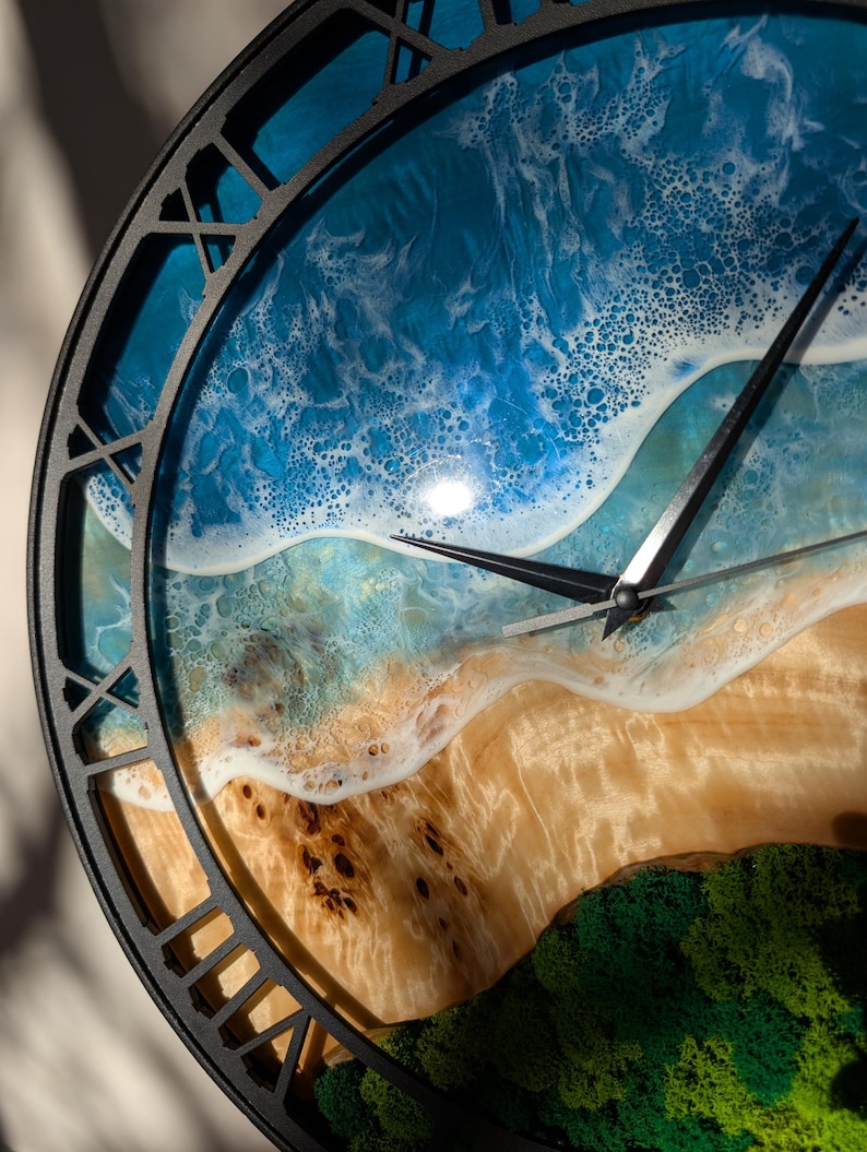 Pu&ograve; includere: Un orologio da parete rotondo con una cornice in metallo nero. Il quadrante dell'orologio presenta una scena di spiaggia con oceano blu, onde bianche e una riva sabbiosa. Le lancette dell'orologio sono nere.