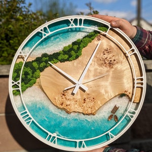 Op de afbeelding: Een witte ronde klok met Romeinse cijfers. De wijzerplaat heeft een kunstig harsontwerp van een strandscène met groen mos, blauw water en witte golven.