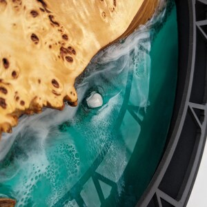 May include: A close-up of a decorative wooden art piece. The piece features a light-colored, textured wood section with a dark teal resin section. A white, shell-like object is embedded in the resin, and the piece is framed by a dark gray border.
