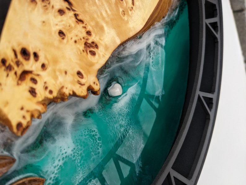May include: A close-up of a decorative wooden art piece. The piece features a light-colored, textured wood section with a dark teal resin section. A white, shell-like object is embedded in the resin, and the piece is framed by a dark gray border.