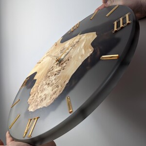 May include: A round wall clock with a dark gray resin border and a light wood grain center. The clock features gold Roman numerals and hands. The wood grain has a unique pattern, adding a natural element to the design.