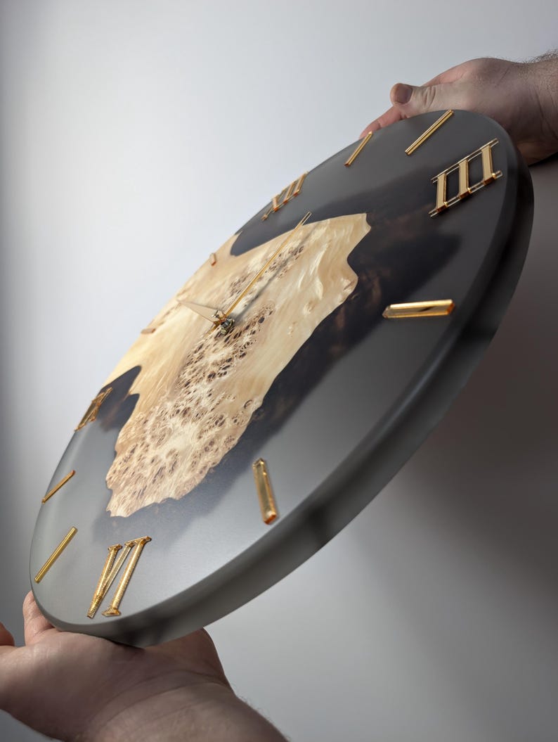 May include: A round wall clock with a dark gray resin border and a light wood grain center. The clock features gold Roman numerals and hands. The wood grain has a unique pattern, adding a natural element to the design.