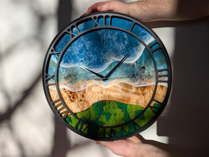Pu&ograve; includere: Orologio da parete rotondo con cornice nera e numeri romani. Il quadrante presenta una scena di spiaggia con onde blu dell'oceano, una riva sabbiosa e una collina verde e muschiosa. Le lancette dell'orologio sono nere.