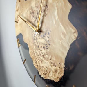 May include: A round wall clock with a light wood grain center and a dark gray resin border. The clock hands and Roman numeral hour markers are gold. The wood grain has a unique, textured pattern.