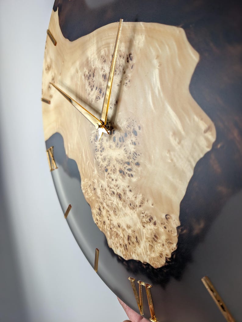May include: A round wall clock with a light wood grain center and a dark gray resin border. The clock hands and Roman numeral hour markers are gold. The wood grain has a unique, textured pattern.