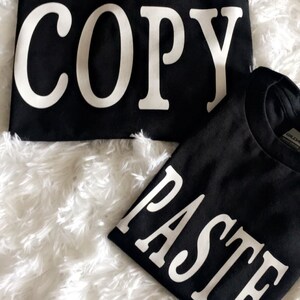 May include: Two black t-shirts with white text. The top shirt says "COPY" and the bottom shirt says "PASTE".