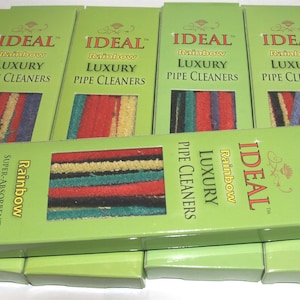 May include: Several boxes of "IDEAL Rainbow Luxury Pipe Cleaners" are stacked. Each box is green with the product name in red and white. The pipe cleaners inside are multi-colored, with stripes of red, yellow, blue, and black. The boxes are labeled "36 Count."
