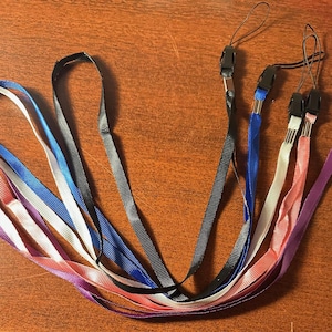 May include: A collection of lanyards in various colors, including purple, pink, white, blue, and black. Each lanyard has a thin, flat strap and a black plastic clasp. The lanyards are arranged on a wooden surface.