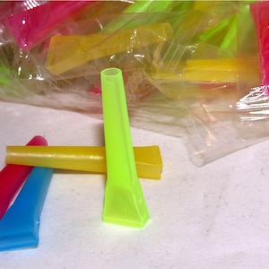 May include: A collection of colorful plastic applicator tips in shades of pink, blue, yellow, and green. Some tips are arranged in a small group, while others are in a clear plastic bag. The tips have a tapered design with a small opening.
