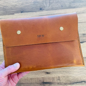 May include: A brown leather laptop sleeve with a flap closure and two brass poppers. The sleeve has the initials "SPW" embossed on the front.