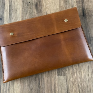 May include: A rectangular brown leather document sleeve featuring a fold-over flap secured by two circular brass-toned metal snap buttons. The edges are finished with visible dark stitching along the sides and bottom of the flat pouch design.