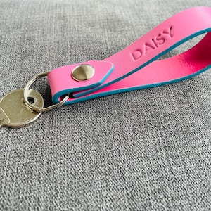 May include: Pink leather keychain with a blue trim and the word "DAISY" embossed on it. The keychain has a silver key ring attached.