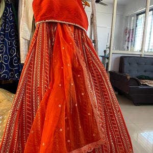 Könnte beinhalten: Ein leuchtend orangefarbenes zweiteiliges Outfit, wahrscheinlich ein traditionelles indisches Kleidungsstück, hängt an einem Gestell. Das Oberteil ist im Neckholder-Stil gehalten, und der Rock weist aufwendige silberne Details auf. Ein durchsichtiger oranger Schal ist über den Rock drapiert.