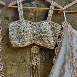 May include: A gold-toned, heavily embellished top with mirror and bead detailing, hanging on a wooden hanger. The top features wide straps and a decorative fringe. A white and gold sheer fabric is visible on the right.