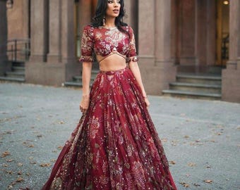 May include: A burgundy two-piece formal outfit with floral embroidery. The top has short sleeves and a cropped design, while the skirt is floor-length. The ensemble is adorned with gold accents and sequins, creating a glamorous look.