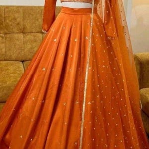 Könnte beinhalten: Ein orangefarbener Satin-Lehenga mit passendem langärmligem Crop-Top und einem transparenten Dupatta. Der Lehenga hat kleine, verstreute weiße Verzierungen und eine goldene Bordüre. Das Outfit wird auf einer Schaufenquin präsentiert.