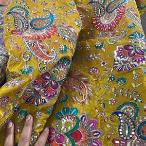 Peut inclure: Tissu jaune moutarde avec broderies complexes repr&eacute;sentant des motifs floraux et cachemire. Le tissu est orn&eacute; de paillettes et de couleurs vives comme le rouge, le bleu et le vert. L'image montre un gros plan de la texture et du motif du tissu.