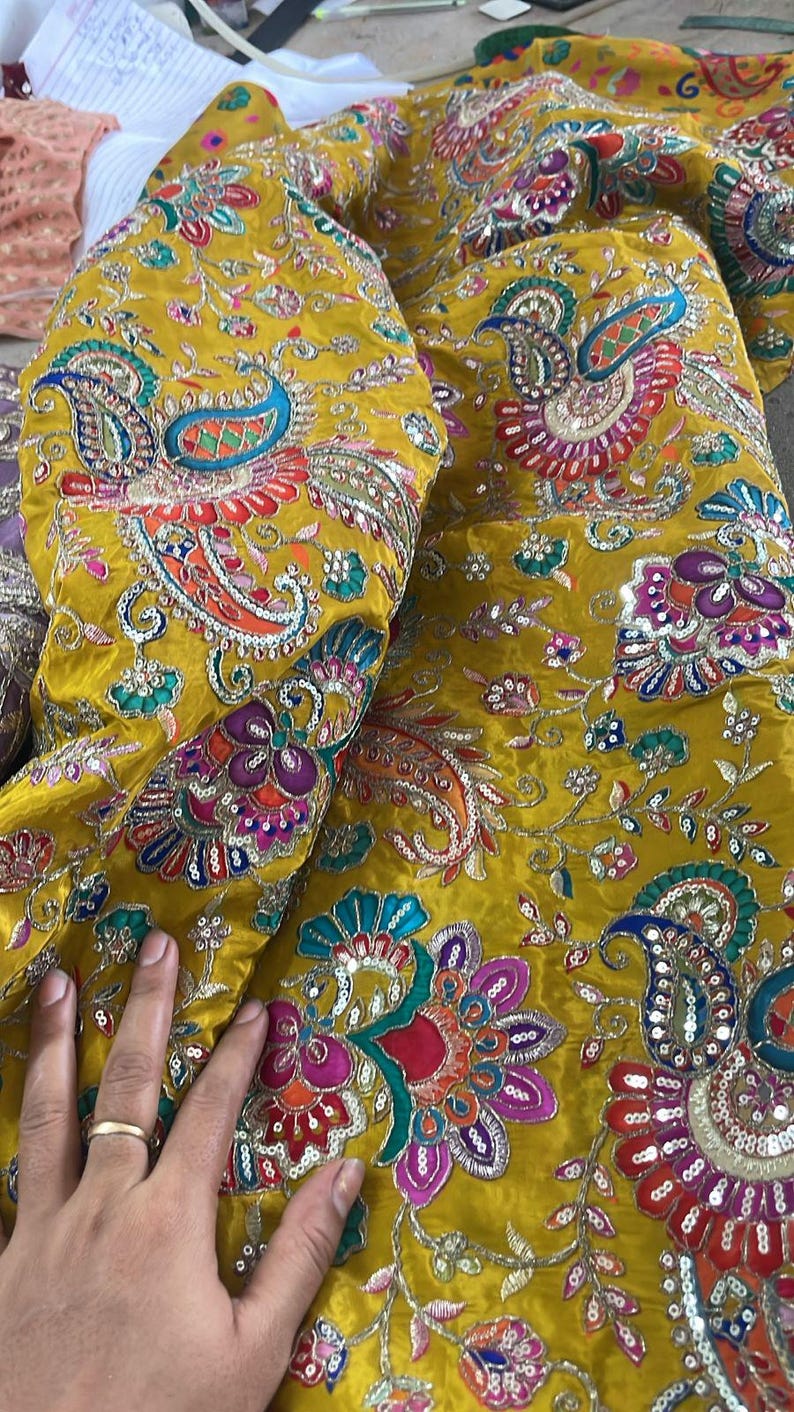 Peut inclure: Tissu jaune moutarde avec broderies complexes repr&eacute;sentant des motifs floraux et cachemire. Le tissu est orn&eacute; de paillettes et de couleurs vives comme le rouge, le bleu et le vert. L'image montre un gros plan de la texture et du motif du tissu.