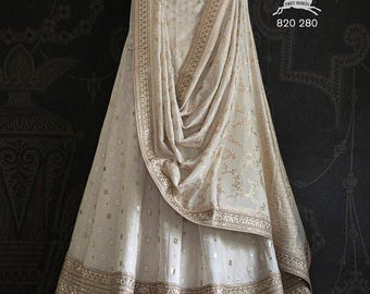 Può includere: Gonna lehenga e dupatta abbinato color avorio, impreziositi da ricami dorati e piccoli dettagli quadrati dorati. Il dupatta è drappeggiato sulla gonna, mostrando un motivo floreale. Il testo "SWATI MANISH 820 280" è visibile.