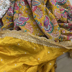 Peut inclure: Tissu jaune moutarde avec broderies et ornements complexes. Le motif pr&eacute;sente des motifs cachemire color&eacute;s et une d&eacute;licate bordure dor&eacute;e. Le tissu est pli&eacute;, r&eacute;v&eacute;lant le savoir-faire et la texture du v&ecirc;tement.