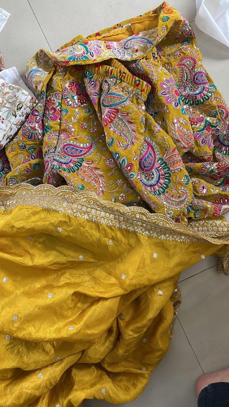 Peut inclure: Tissu jaune moutarde avec broderies et ornements complexes. Le motif pr&eacute;sente des motifs cachemire color&eacute;s et une d&eacute;licate bordure dor&eacute;e. Le tissu est pli&eacute;, r&eacute;v&eacute;lant le savoir-faire et la texture du v&ecirc;tement.