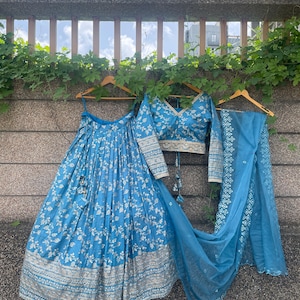 May include: A three-piece blue floral embroidered outfit, including a long skirt, a long-sleeved top, and a matching scarf. The skirt and top are adorned with white floral patterns and silver embroidery. The outfit is hanging on wooden hangers against a textured wall.