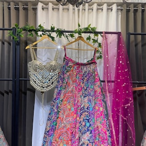 Puede incluir: Un vibrante conjunto lehenga multicolor con un top espejado y una falda estampada. La falda presenta un diseño de cachemira en tonos rojos, azules y morados. Una dupatta rosa transparente con lentejuelas doradas cuelga a un lado.