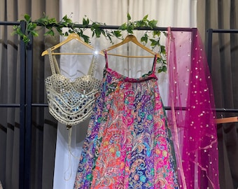 May include: A vibrant, multi-colored lehenga set with a mirrored top and a patterned skirt. The skirt features a paisley design in shades of red, blue, and purple. A sheer pink dupatta with gold sequins hangs to the side.
