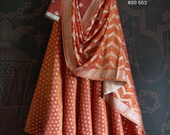 Può includere: Un lehenga in seta color ruggine con un motivo ricamato in argento. Il lehenga è abbinato a una blusa in seta color ruggine abbinata con ricami argentati. Il tessuto ha una lucentezza sottile e un delicato motivo floreale. Il lehenga è esposto su un appendiabiti su uno sfondo scuro. Il testo "SWATI MANISH 820 552" è visibile nell'angolo in alto a destra dell'immagine.