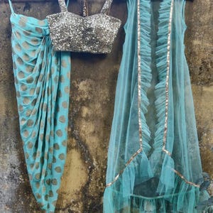 May include: A three-piece outfit comprising a turquoise saree with gold polka dots, a silver sequined bustier top, and a turquoise tulle jacket with ruffled trim and gold accents. The garments are displayed on wooden hangers.