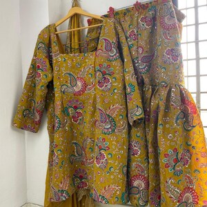 Peut inclure: Ensemble jaune moutarde compos&eacute; d'un haut et d'un pantalon &eacute;vas&eacute;, orn&eacute; d'un motif cachemire rose, vert et bleu. La tenue comprend une &eacute;charpe assortie avec une bordure dor&eacute;e, le tout suspendu &agrave; un cintre en bois et des pinces. Le tissu a un aspect textur&eacute;.