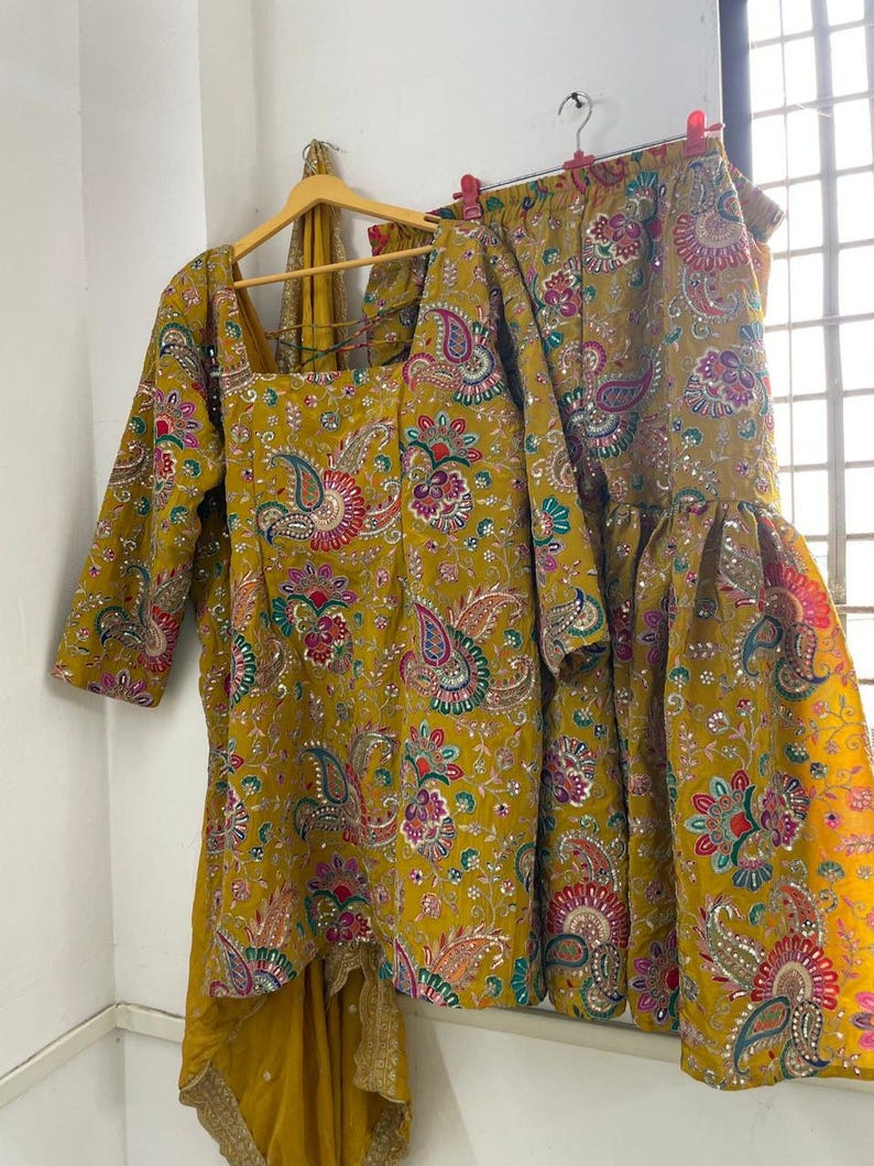 Peut inclure: Ensemble jaune moutarde compos&eacute; d'un haut et d'un pantalon &eacute;vas&eacute;, orn&eacute; d'un motif cachemire rose, vert et bleu. La tenue comprend une &eacute;charpe assortie avec une bordure dor&eacute;e, le tout suspendu &agrave; un cintre en bois et des pinces. Le tissu a un aspect textur&eacute;.