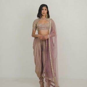 May include: A three-piece ensemble featuring a beige cropped blouse with intricate embroidery and a keyhole neckline, paired with matching beige dhoti-style trousers and a sheer mauve dupatta with a gold embroidered border along the edges.