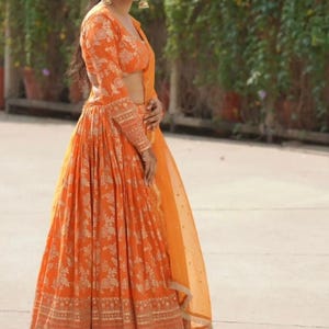 Könnte beinhalten: Ein orangefarbenes Lehenga-Choli-Ensemble mit floralem Muster. Das Outfit besteht aus einer langärmeligen Bluse und einem fließenden Rock, beide mit goldenen Blumenmustern verziert. Ein durchsichtiges orangefarbenes Dupatta vervollständigt den Look. Der Text "Rudra Fashion Boutique" ist sichtbar.