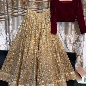Puede incluir: Una falda dorada con lentejuelas y un top granate de manga larga cuelgan de perchas de madera. La falda está muy adornada con lentejuelas y tiene un borde decorativo. La parte superior está hecha de un material similar al terciopelo. El texto "LABEL LUSH by Sabina Khan" es visible.