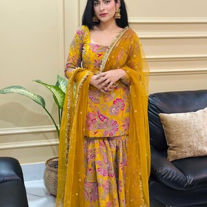 Peut inclure: Une femme porte un salwar kameez jaune vif avec un dupatta assorti. La tenue pr&eacute;sente un motif floral cachemire rose et violet. Des bordures dor&eacute;es ornent les bords du dupatta et l'encolure du haut. La femme porte &eacute;galement des boucles d'oreilles dor&eacute;es.