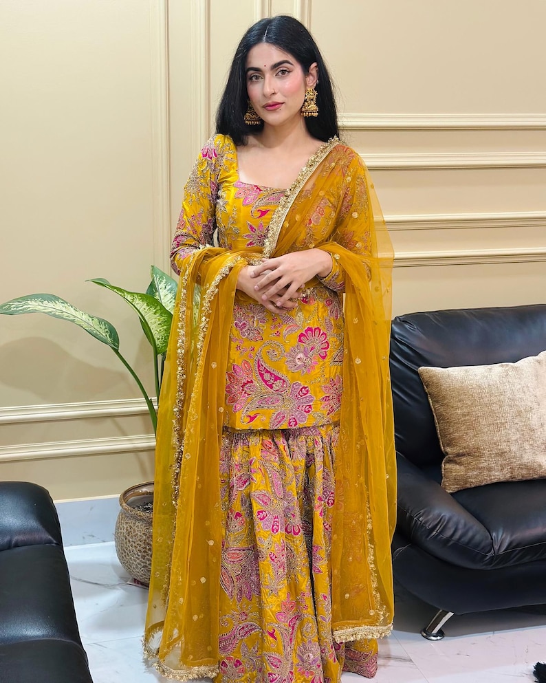 Peut inclure: Une femme porte un salwar kameez jaune vif avec un dupatta assorti. La tenue pr&eacute;sente un motif floral cachemire rose et violet. Des bordures dor&eacute;es ornent les bords du dupatta et l'encolure du haut. La femme porte &eacute;galement des boucles d'oreilles dor&eacute;es.