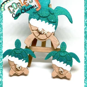 May include: Three teal green glitter sea turtle car charms with white foam and a seashell on a sandy brown base.  Cobles Car Charms is written on the sign above the largest turtle.