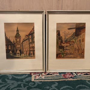 May include: Two framed watercolor paintings. The left painting depicts a European city scene with a clock tower. The right painting shows a rural landscape with mountains. Both paintings are in silver frames with white mats.