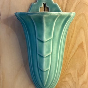 May include: A turquoise ceramic wall pocket vase with a decorative top and a flared, fluted body. The vase has a cross-shaped opening at the top for hanging. The vase is mounted on a light-colored wooden surface.