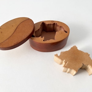May include: A wooden puzzle box with a lid shaped like a bean. The box is open and the puzzle piece is removed, revealing the shape of the piece inside the box.