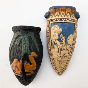 May include: Two vintage ceramic wall vases. One vase is black with a green palm tree and an orange camel. The other vase is blue and white with a knight riding a horse.
