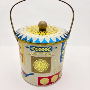 May include: A cylindrical metal tin with a beige base color, featuring a gold-toned wire handle and a removable lid with a central knob. The exterior is decorated with colorful graphic patterns including blue, red, and yellow geometric shapes.